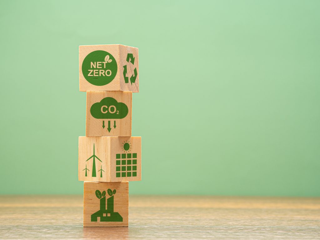 A green background featuring wooden building blocks on a wooden surface. Building blocks feature green images of Net Zero related items such as wind turbines and recycling symbols.